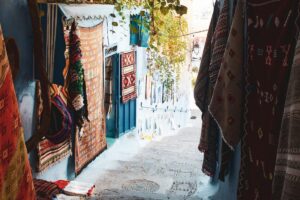 Image d&rsquo;une ruelle étroite dans un endroit probablement marocain, avec des murs bleus et de nombreux tapis colorés suspendus. Les feuilles de plantes poussent au-dessus de la ruelle, donnant un air charmant. Cette image est liée à &ldquo;À propos de BaoLiba – Connecter les influenceurs et les marques marocaines au monde entier&rdquo;, montrant la richesse culturelle marocaine qui peut être mise en valeur par les influenceurs et les marques sur la scène internationale.