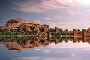 Image présentant un village traditionnel marocain avec des bâtiments en terre cuite, niché au pied d&rsquo;une colline et reflété dans une étendue d&rsquo;eau calme. Les palmiers environnent le lieu, et le ciel est orné de nuages. Cette image est associée aux &ldquo;Questions fréquentes sur BaoLiba et les collaborations d’influence au Maroc&rdquo;, suggérant que BaoLiba s&rsquo;intéresse aux richesses locales et aide à élucider les interrogations autour des partenariats d&rsquo;influence dans ce pays aux paysages emblématiques.