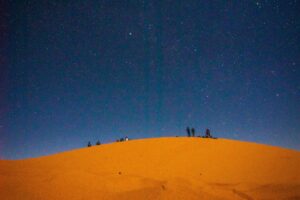 Image montrant une dune de sable sous un ciel étoilé. Plusieurs personnes se trouvent sur la dune, peut - être à profiter de la beauté du paysage nocturne. Cette image est associée aux &ldquo;Tendances du marketing sur les réseaux sociaux au Maroc en 2025&rdquo;, suggérant que le Maroc, avec ses paysages uniques comme celui - ci, offre de nombreuses opportunités pour le marketing sur les réseaux sociaux, en utilisant l&rsquo;attrait naturel du territoire.